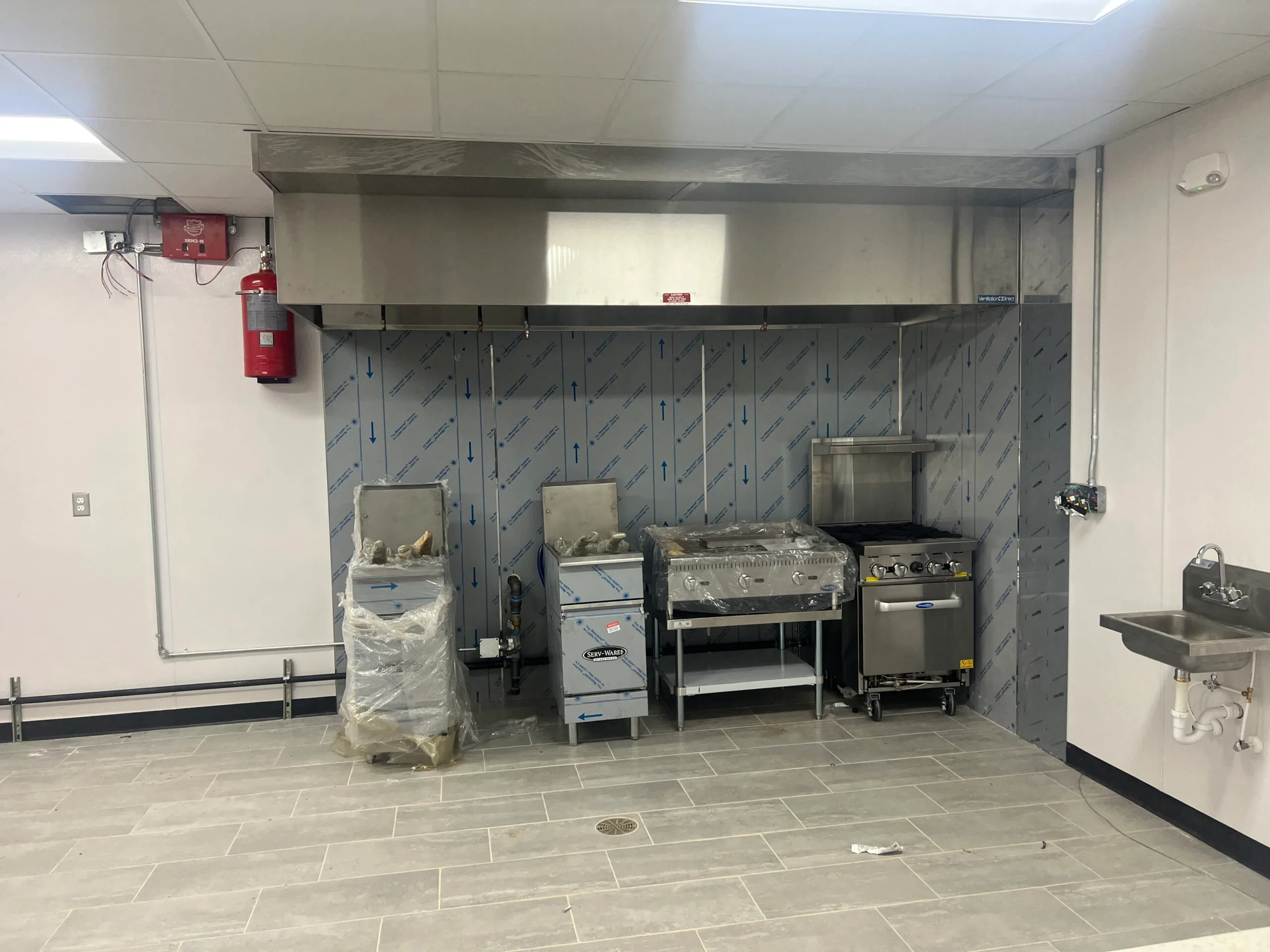A commercial kitchen with new, unpacked appliances, including a fryer, grill, and range under a hood. Nearby are a wall-mounted sink and fire extinguisher. The space also features efficient HVAC for optimal air conditioning.
