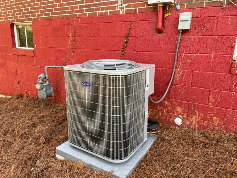 Air Conditioning Services in Sandy Springs 4 An expertly done AC installation features an outdoor air conditioning unit on a concrete slab near a red brick wall, surrounded by dry pine needles.