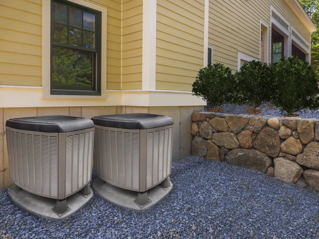 Two air conditioning units sit on a gravel surface by the yellow exterior of a house, bordered by a stone wall and bushes. For optimal performance, consult HVAC contractors in Georgia to ensure everything runs smoothly.