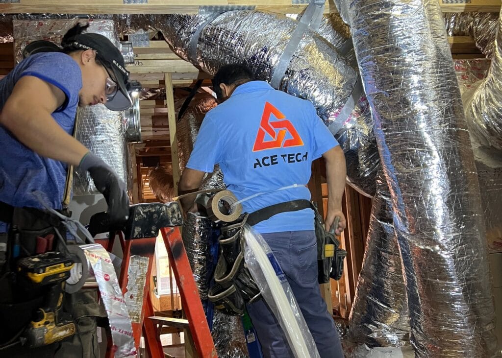 Two workers, renowned HVAC contractors in Georgia, install ductwork at a construction site. One wears a blue shirt with "Ace Tech" on the back, surrounded by an array of tools.