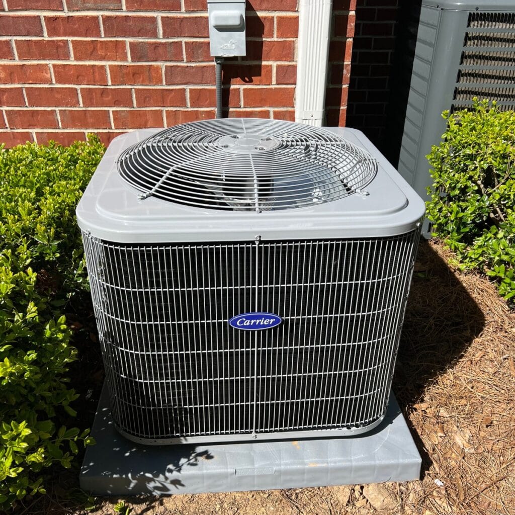 Outdoor air conditioning unit with a metal grate on top, installed by skilled HVAC contractors in Georgia, nestled next to a brick wall and surrounded by lush greenery.