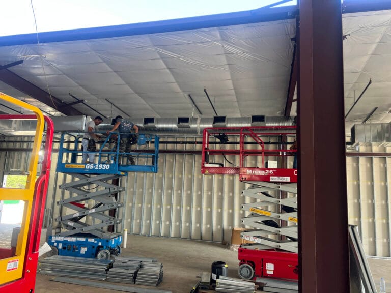 In a large unfinished building, two workers on scissor lifts meticulously install ductwork, their teamwork as seamless as a dynamic content widget integrating into a webpage.