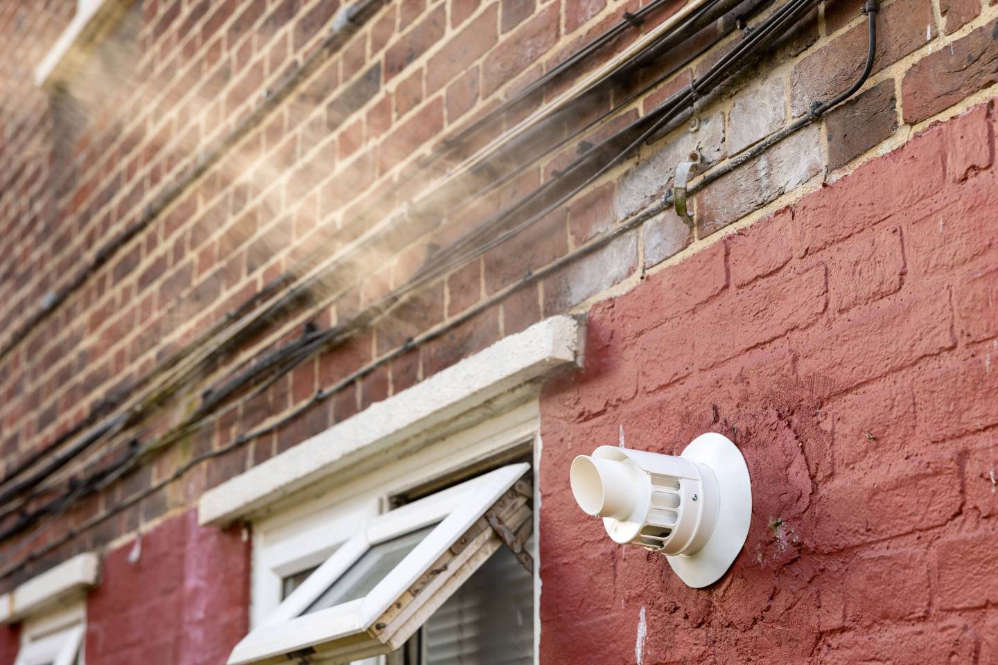 Atlanta Air Quality Services 6 A vent, freshly serviced with air duct cleaning, is installed on a red brick wall below electrical wires, next to open windows. Sunlight shines across the image.
