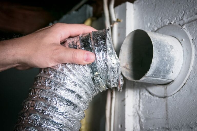 Atlanta Air Quality Services 4 A hand is carefully holding a flexible silver duct near a round wall vent opening in a building, ready for dryer vent cleaning.