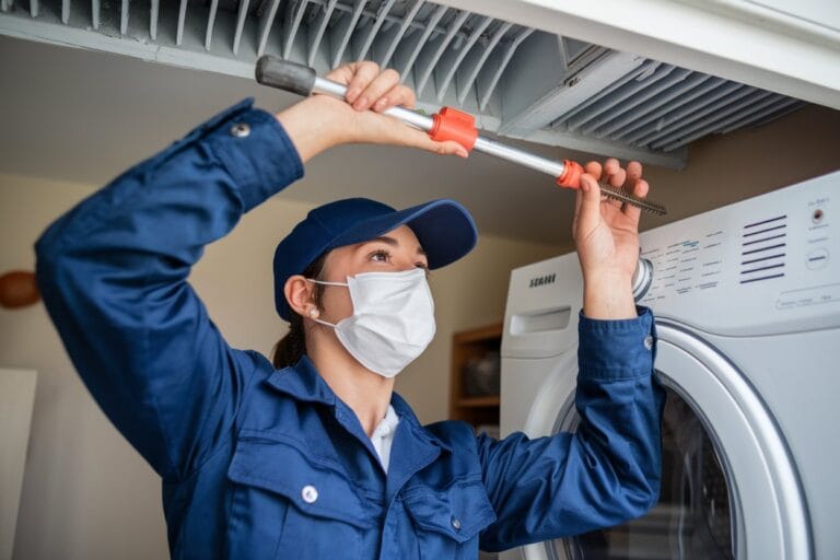 A person in a blue uniform and mask is meticulously cleaning a dryer vent with a brush, ensuring optimal airflow. Nearby, the washing machine stands ready for service. This dynamic content scene could enhance any home care guide or widget-driven interface.