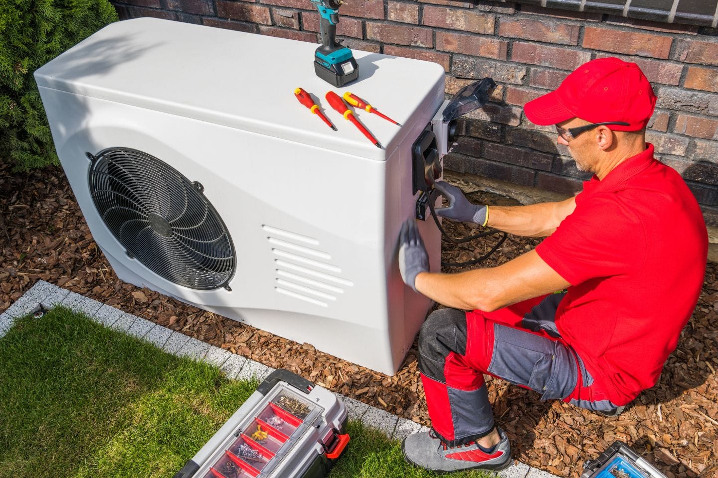 A technician in a red uniform and gloves diligently works on an outdoor air conditioning unit, with a variety of tools and a widget atop the unit, while a toolbox rests on the grass.