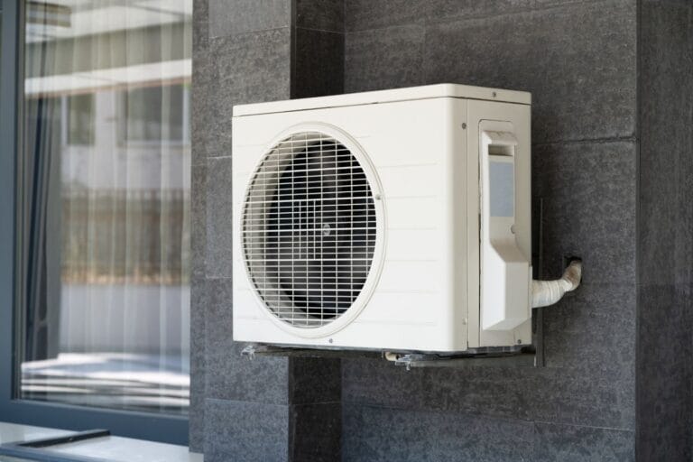 A sleek outdoor heat pump is mounted on a dark gray wall next to a window, efficiently regulating temperatures and providing optimal comfort.