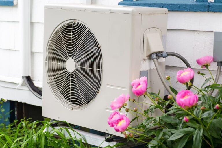 An outdoor heat pump is mounted on a wooden wall, surrounded by blooming pink flowers and greenery.