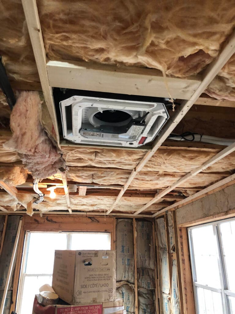 Room under construction with exposed insulation and an uninstalled ceiling fan. Boxes are stacked near the windows, awaiting AC installation to complete the setup efficiently.