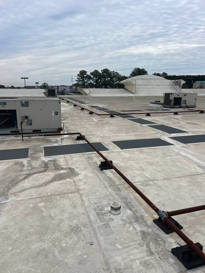 A flat commercial rooftop dotted with multiple HVAC units, vent pipes, and conduit lines lies beneath a cloudy sky, framed by trees in the background. This setup exemplifies expert HVAC services ensuring optimal air conditioning installation.
