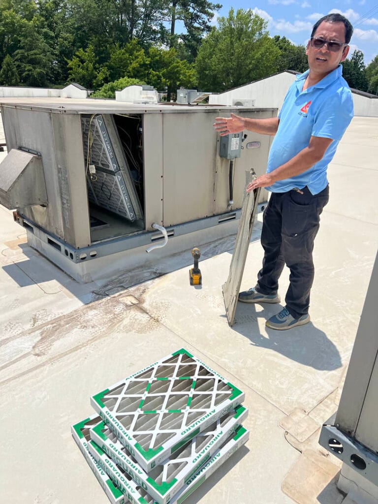 A person stands on a rooftop next to an HVAC unit, holding an air filter. Several replacement filters and a drill are on the ground. Trees and buildings grace the background, setting the scene for AC installation maintenance.