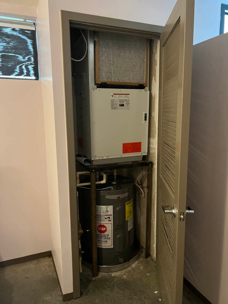 A closet containing a water heater and an efficient HVAC unit with visible pipes and ductwork, showcasing expert AC installation. The door is partially open.
