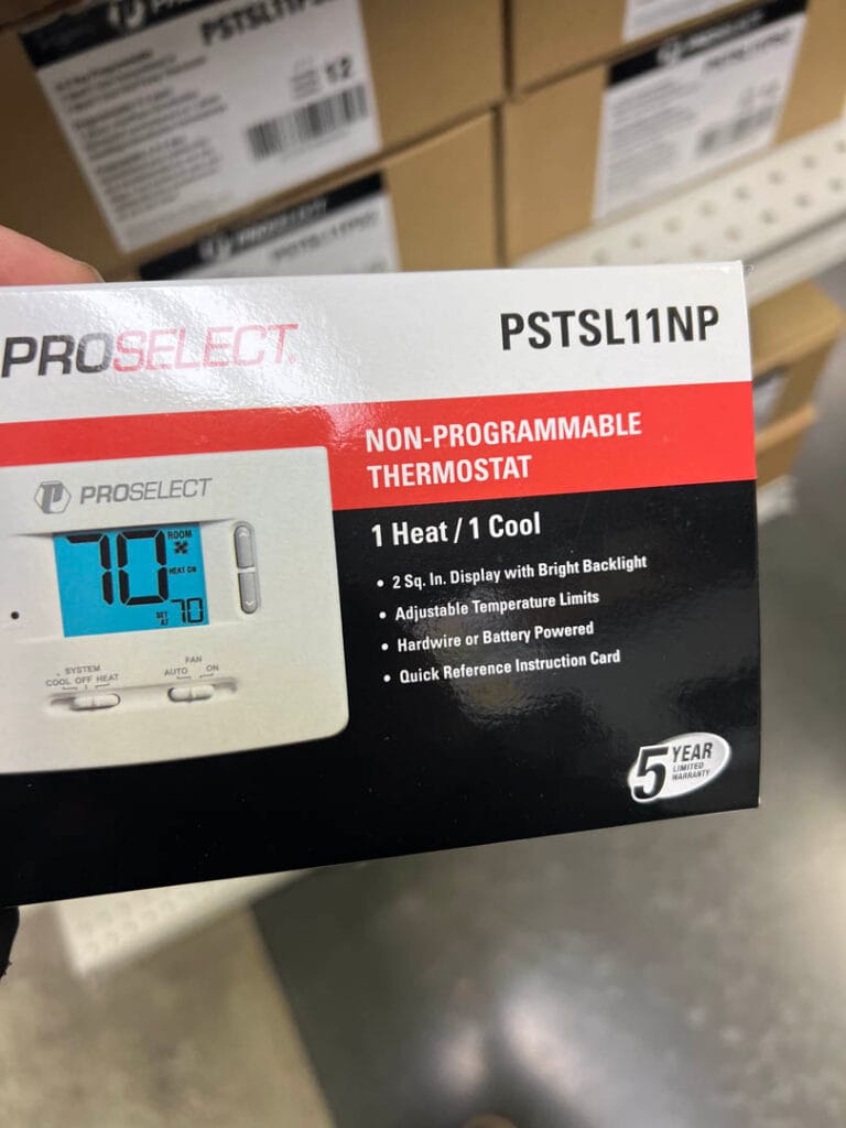 A person holds a box of a ProSelect PSTSL11NP non-programmable thermostat with features like 1 heat/1 cool function and a 5-year warranty—an ideal addition to your next AC installation project.