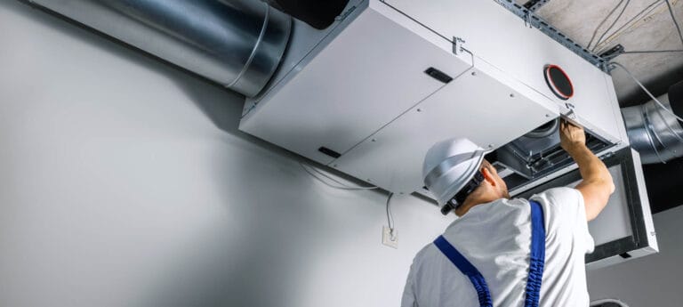 Technician in a white hard hat and blue overalls inspects a ceiling-mounted HVAC duct system.