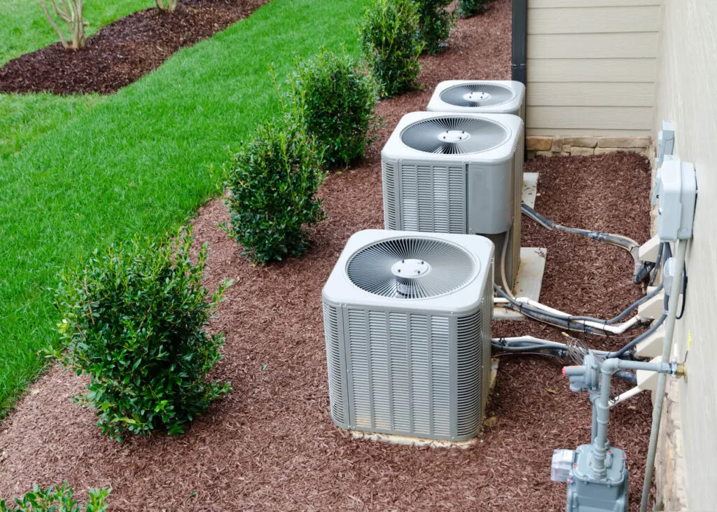 About 4 Three outdoor air conditioning units installed beside a house on mulch, with small bushes and green grass nearby.
