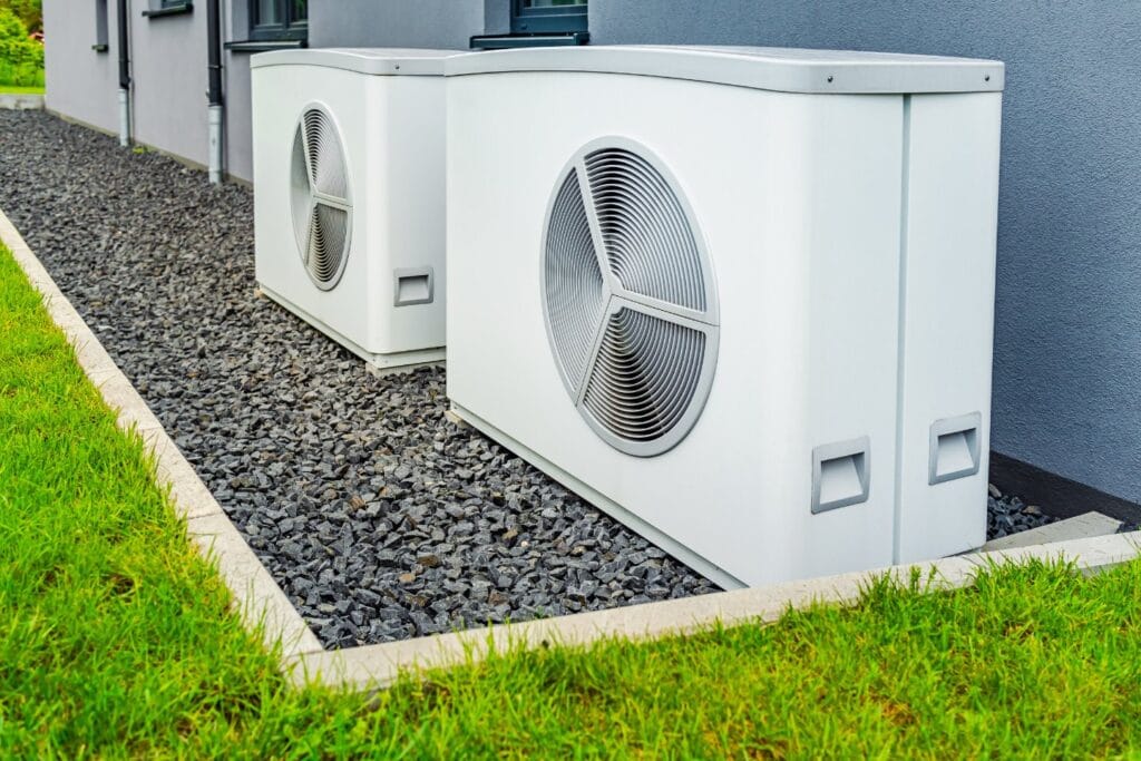 Two white outdoor air conditioning units, freshly installed, rest on a bed of gray gravel beside a building. A lush grassy area stretches out in the foreground, framing the scene of this meticulous AC installation.