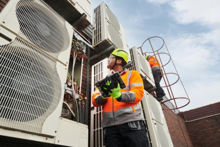 Two workers in safety gear inspect rooftop HVAC units. One holds tools, while the other climbs a ladder to ensure proper AC installation. The sky forms a clear backdrop for their meticulous task.