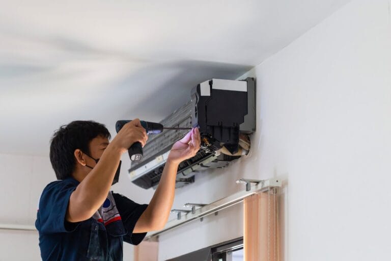 A person in a dark shirt is using a power drill for an HVAC repair on an air conditioning unit mounted on a wall.