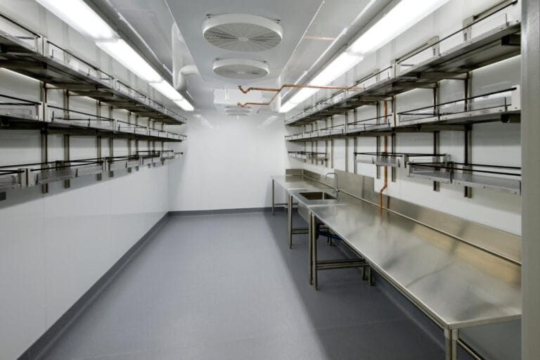 Empty commercial stainless steel kitchen with shelves, counters, and overhead lighting, featuring a gray floor and white walls. The space is ready for setup with state-of-the-art refrigeration services to meet all culinary needs efficiently.
