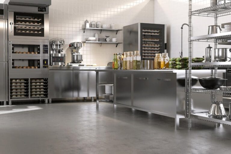 The industrial kitchen boasts stainless steel counters, shelves, and state-of-the-art refrigeration equipment. Ovens piled with trays flank mixers on the counter, while bottles and vegetables are prominently displayed.