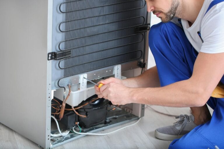 A technician in blue overalls expertly provides refrigeration services, using a screwdriver to repair the back of a refrigerator on a wooden floor.