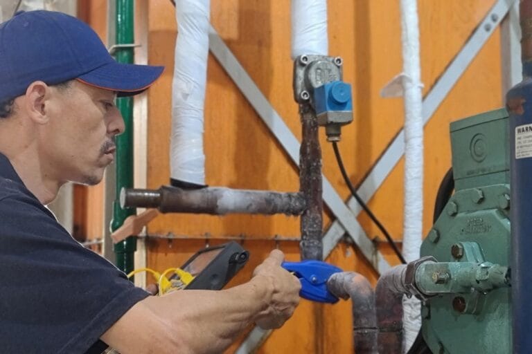 A person in a blue cap, providing refrigeration services, diligently inspects industrial pipes in a facility with orange walls and wrapped insulation.