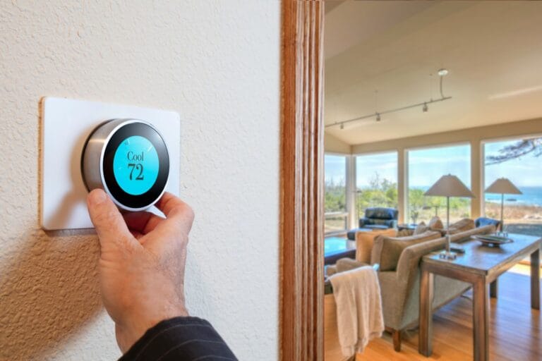 A hand adjusts a smart thermostat widget set to 72 degrees on a wall. In the background, there is a living room with large windows and dynamic content featuring an ocean view.