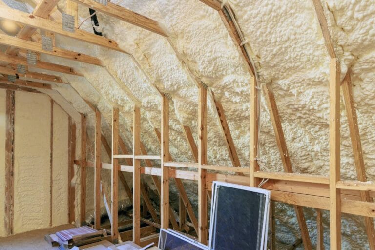 Attic under renovation with exposed wooden framing and spray foam insulation on walls and ceiling, while panels and building materials lie scattered on the floor, awaiting transformation into a dynamic content widget of comfort.
