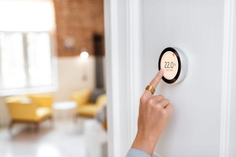 A person adjusts a smart thermostat on a wall, displaying 22.0°C. A sunlit living room with yellow chairs and a dynamic content widget enhancing the ambiance is visible in the background.