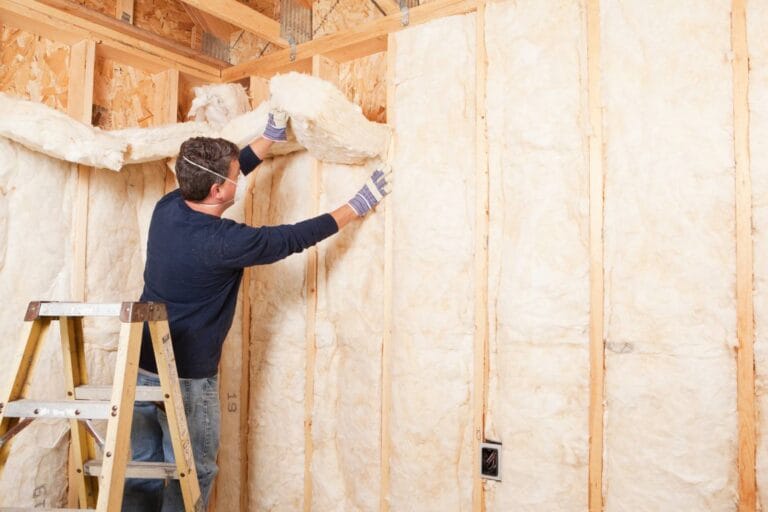 In a partially constructed building, a person in a blue shirt stands on a ladder, skillfully installing fiberglass insulation into the wall cavity. With gloves on for safety, this dynamic content showcases the process seamlessly.