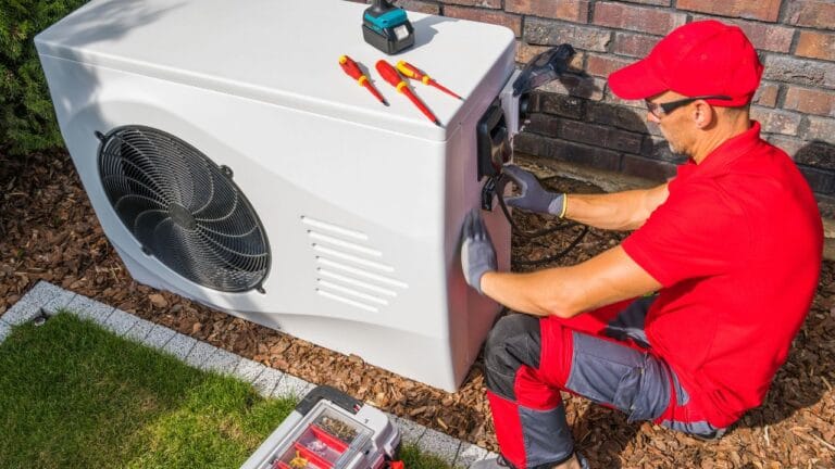 AC Repair & Maintenance 5 A technician in a red uniform and gloves is expertly repairing a large white outdoor HVAC unit near a brick wall, tools scattered nearby—a testament to the reliable Heat Pump Services in Georgia.
