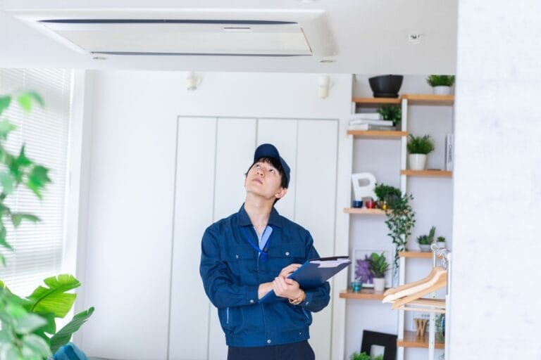 A technician in a crisp blue uniform inspects a ceiling air conditioning unit, diligently holding a clipboard. Specializing in HVAC services, their expert eye ensures every detail is meticulously checked to maintain optimal performance.