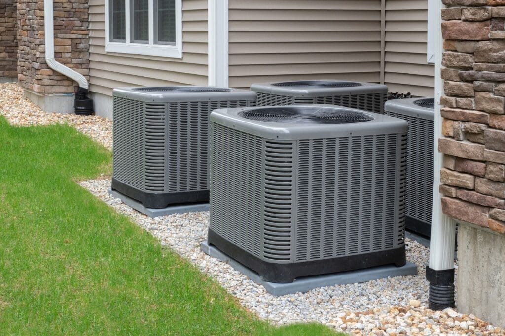 How To Decrease Energy Bills: Four gray outdoor air conditioning units sit on a bed of white stones beside a house with beige siding and brick details, seamlessly blending into their green grass surroundings. As vital components of our home's comfort, these units underscore the importance of reliable air conditioning services to save on energy bills.