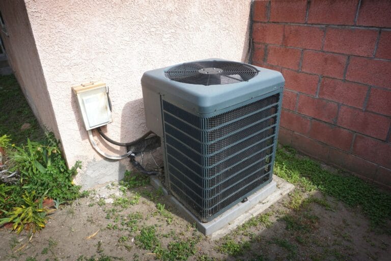 An outdoor air conditioning unit, expertly installed next to a building wall on a concrete pad, is surrounded by grass and a small bush. This setup enhances the efficiency of cooling services provided to the building occupants.