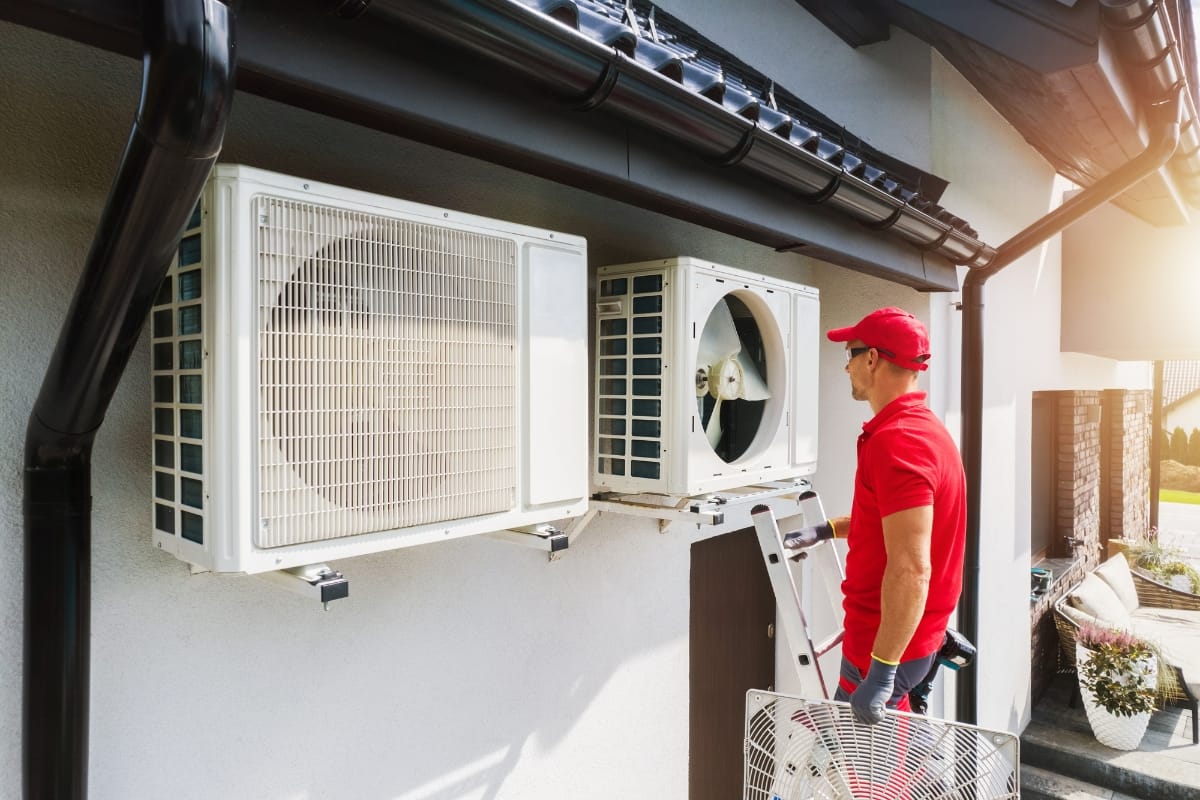 A technician in a red uniform stands on a ladder inspecting and repairing outdoor air conditioning units mounted on the side of a building for essential summer HVAC prep.