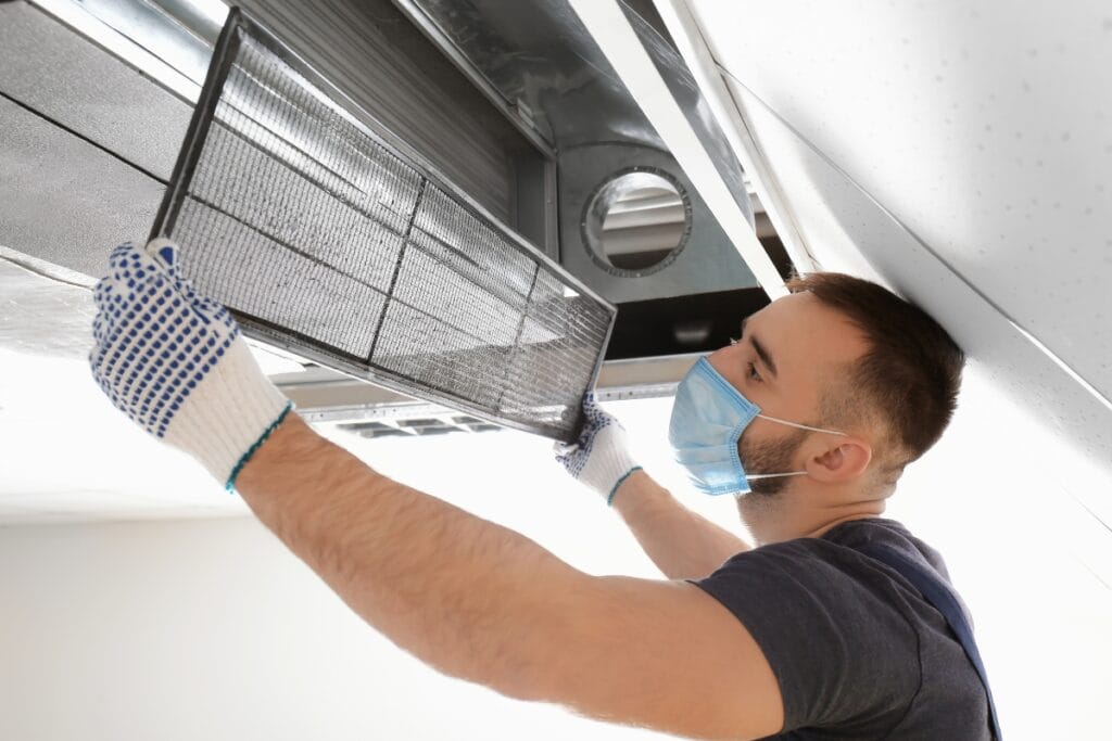 Summer HVAC Prep: How to Get Your System Ready for the Heat 2 A man wearing gloves and a face mask installs or removes an air filter from a ceiling ventilation duct as part of summer HVAC prep.