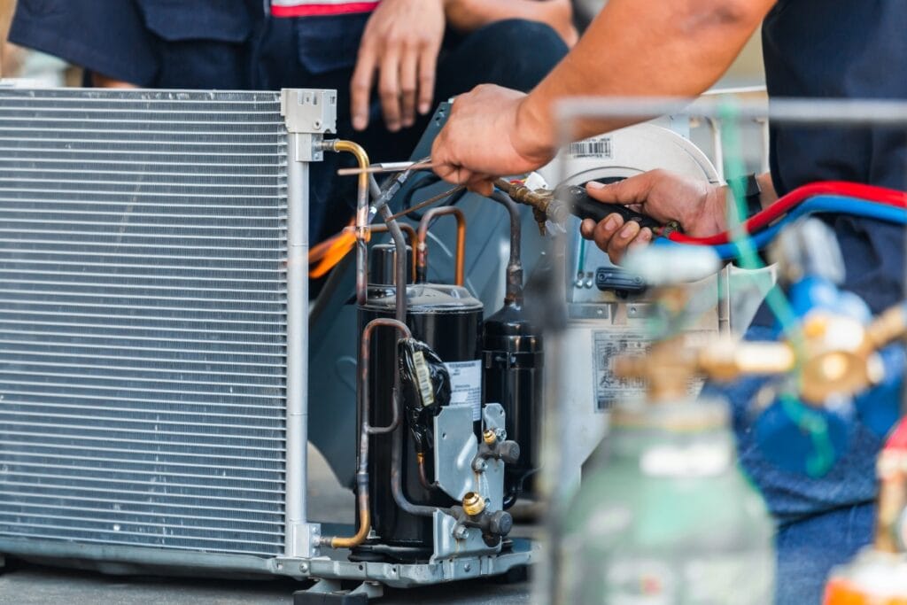 Summer HVAC Prep: How to Get Your System Ready for the Heat 4 Technicians work on air conditioning unit components, connecting wires and tools for summer HVAC prep, with refrigerant tanks visible in the foreground.