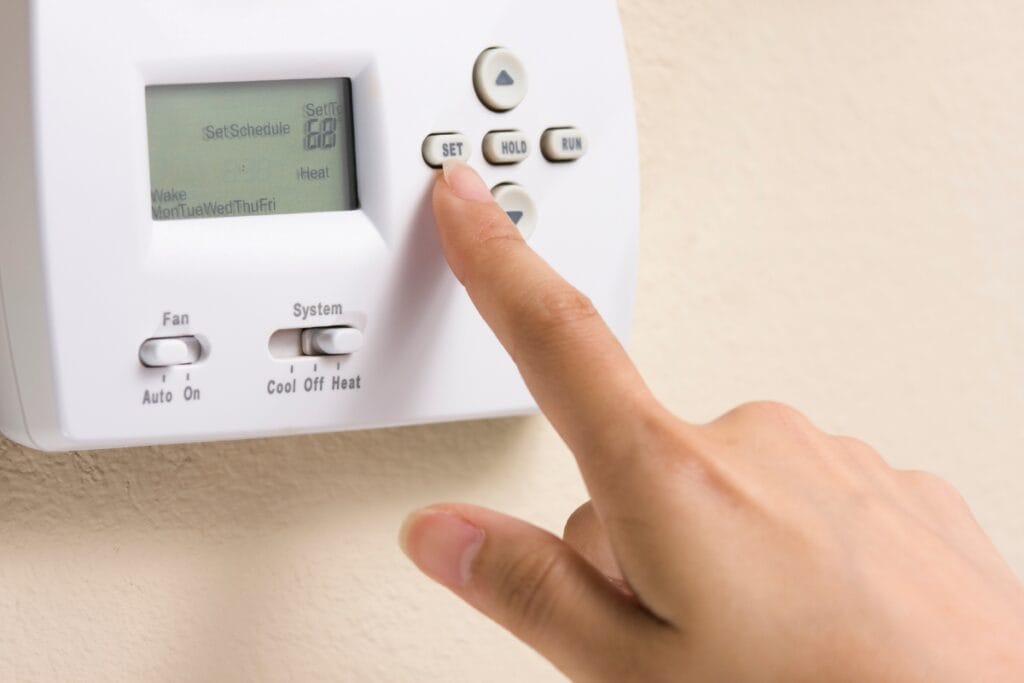 Summer HVAC Prep: How to Get Your System Ready for the Heat 5 A hand pressing the "Set" button on a digital thermostat mounted on a beige wall, perfect for summer HVAC prep.