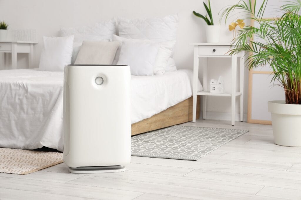 A white air purifier, ideal for air purifiers for allergies, stands on a light wooden floor in a bright, modern bedroom with a bed, bedside table, and green plants.