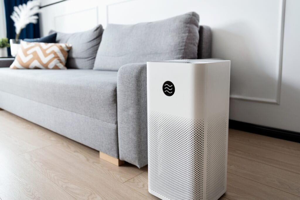 Best Air Purifiers for Allergies: Improve HVAC Air Quality 1 A white air purifier with a digital display, designed as one of the best air purifiers for allergies, stands on a wooden floor in front of a gray sofa in a modern living room.