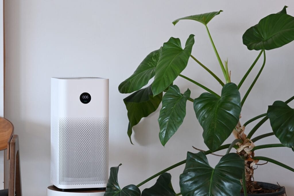 Best Air Purifiers for Allergies: Improve HVAC Air Quality 4 A white air purifier, ideal for air purifiers for allergies, stands next to a large green houseplant with broad leaves against a plain light-colored wall.