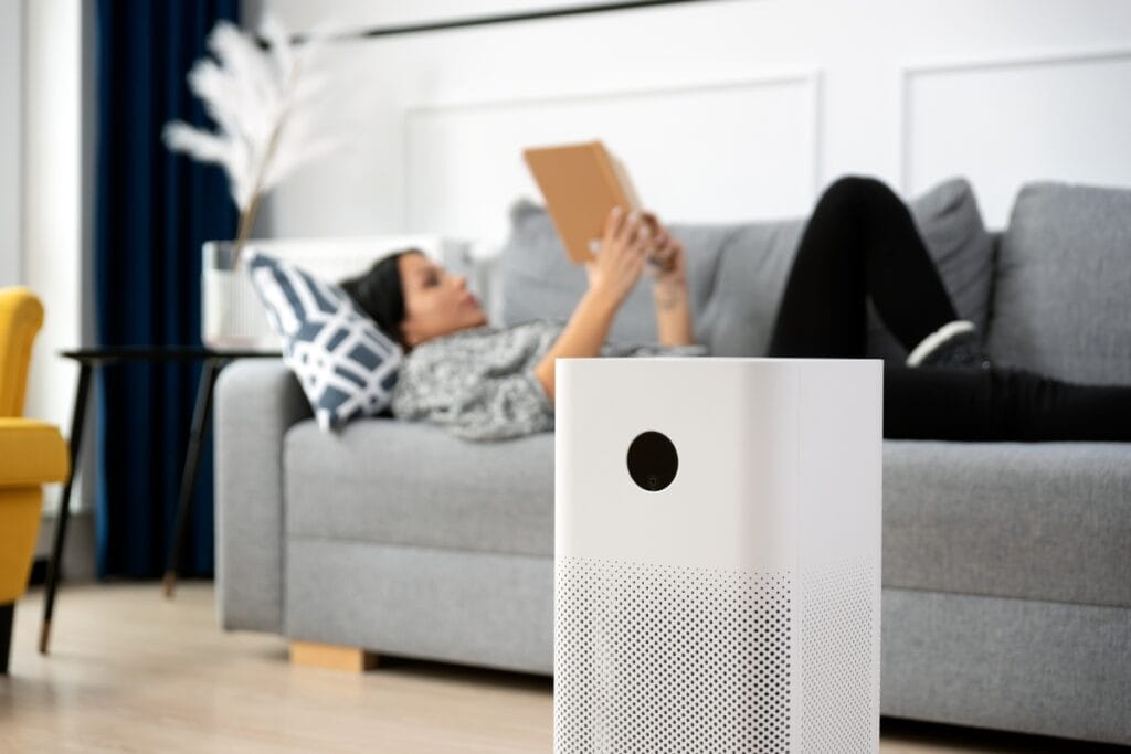Best Air Purifiers for Allergies: Improve HVAC Air Quality 5 A white air purifier, perfect for those seeking air purifiers for allergies, is in the foreground while a person relaxes on a gray sofa in the background, reading a tablet in a modern living room.