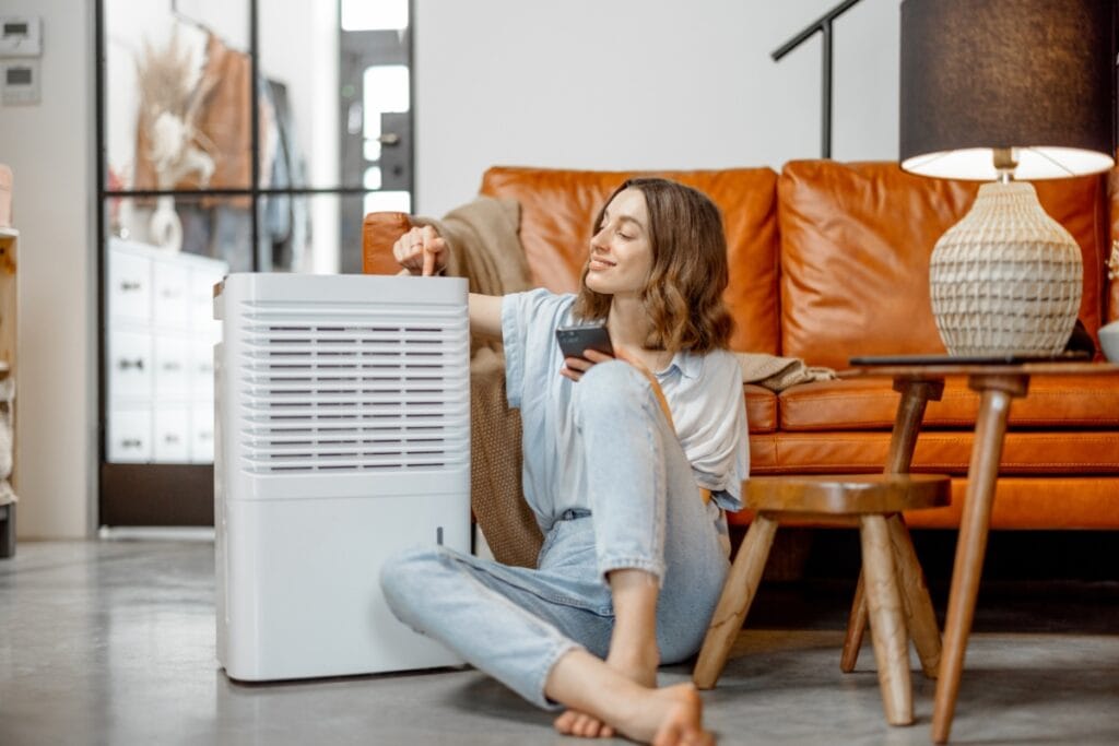 Best Air Purifiers for Allergies: Improve HVAC Air Quality 6 Woman sitting on the floor in a living room, adjusting a dehumidifier next to a brown leather sofa and holding a remote—an ideal setup for those using air purifiers for allergies.