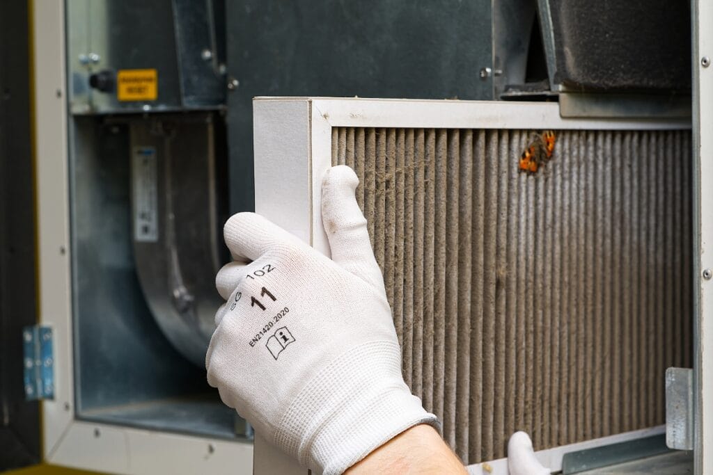 Best Air Purifiers for Allergies: Improve HVAC Air Quality 7 A person wearing white gloves removes a dirty air filter from a ventilation system, an important step in maintaining air purifiers for allergies to ensure cleaner, healthier indoor air.