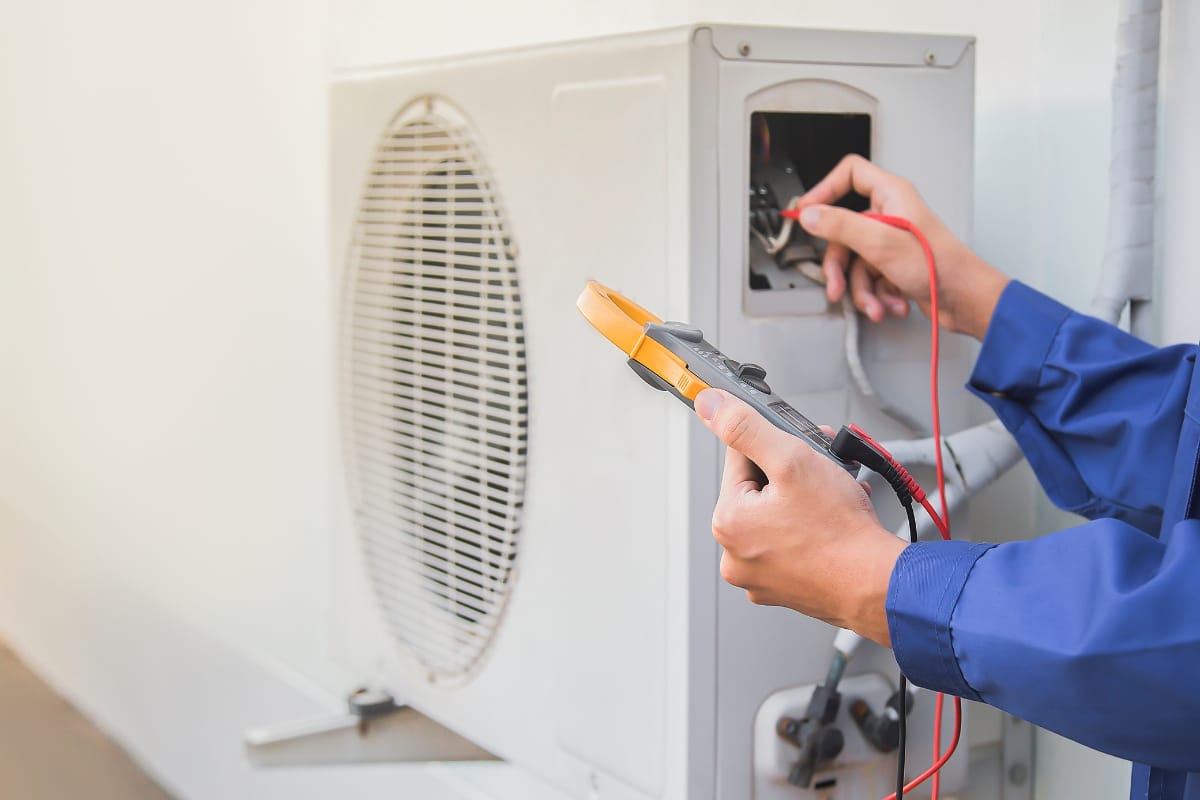 A Commercial HVAC Services In Sandy Springs technician in blue uniform using a digital multimeter to check electrical connections on an outdoor air conditioning unit, ensuring optimal SEER rating HVAC system performance.