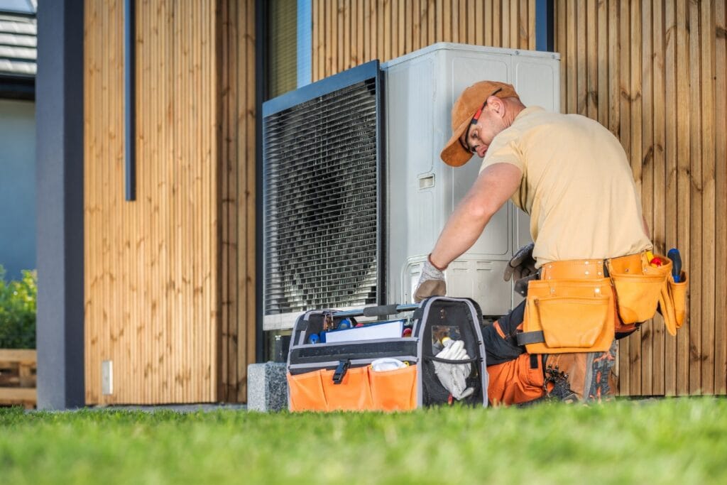 The Ultimate SEER Rating HVAC Guide: Why It’s Crucial for Efficiency 6 A maintenance worker kneels on grass while repairing an outdoor HVAC unit with a high SEER rating next to a building, surrounded by a tool bag and various tools.