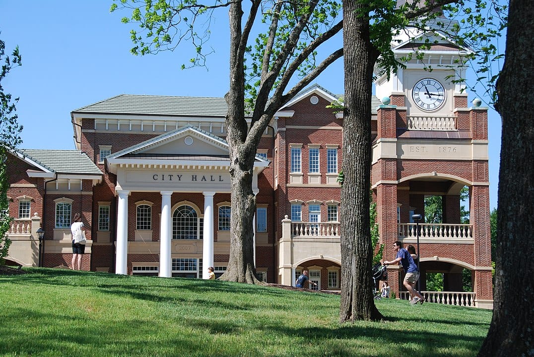 A red-brick city hall building with columns and a clock tower, surrounded by trees and people walking on a grassy lawn, stands as a landmark near trusted Air Conditioning Services in Duluth.