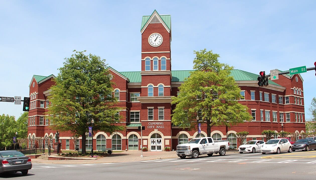 Red brick city hall building with a green roof and clock tower, located at a busy intersection in Cumming, where cars and trees surround the area—an iconic spot near trusted Air Conditioning Services in Cumming.