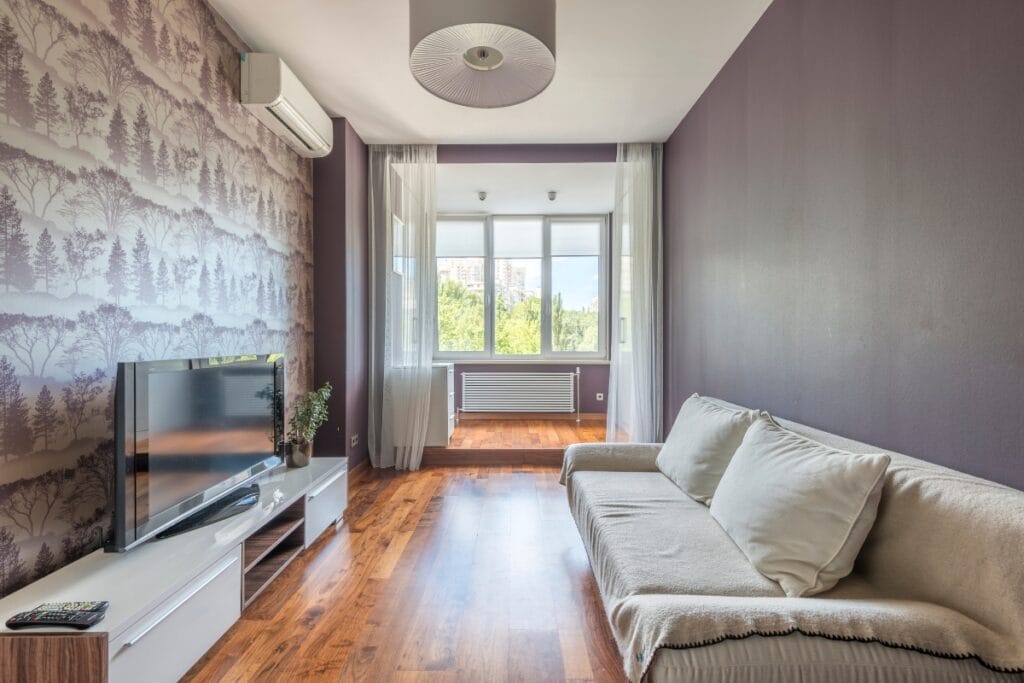 Ductless HVAC Systems: Pros and Cons of Mini-Splits 7 A modern living room with purple walls, a gray sofa, a TV on a white stand, wood flooring, and a large window letting in natural light features comfort enhanced by an efficient ductless HVAC system.