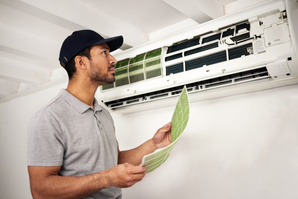 Ductless HVAC Systems: Pros and Cons of Mini-Splits 11 A technician in a gray polo and cap inspects a wall-mounted ductless HVAC unit while holding two green filters.